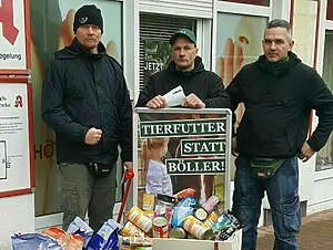 Drei Personen stehen hinter einem Haufen Tierfutter und einem Schild mit der Aufschrift "Tierfutter statt Böller". Sie tragen Kleidung der Partei "Der III. Weg"