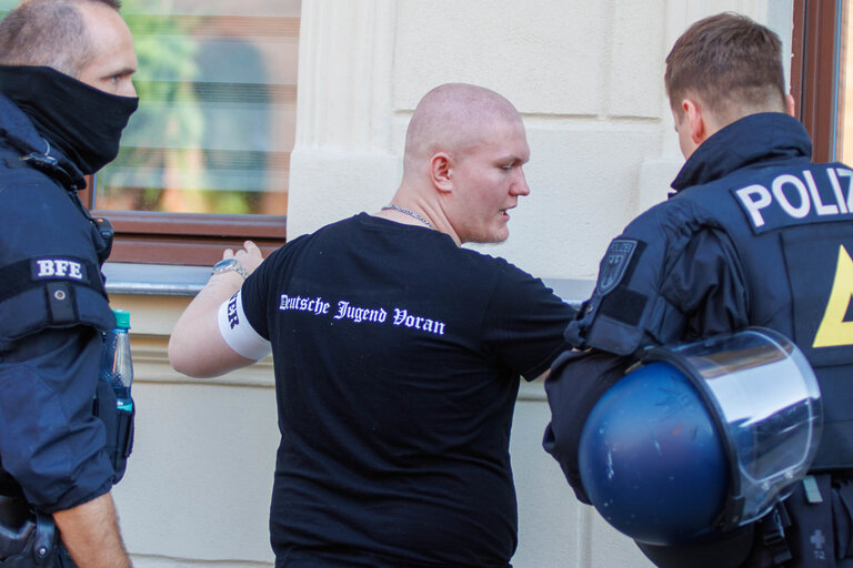 Polizeikontrolle einer glatzköpfigen Person. Auf dessen T-Shirt steht "Deutsche Jugend Voran". Am Arm trägt er eine Binde mit der Aufschrift "Ordner".